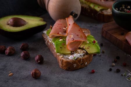 Avocado sandwich close up on gray stone kitchen table with ingredients in the background. Healthy breakfast, healthy food concept. Making delicious avocado toast with nuts, sprouts, ham and spicesの写真素材