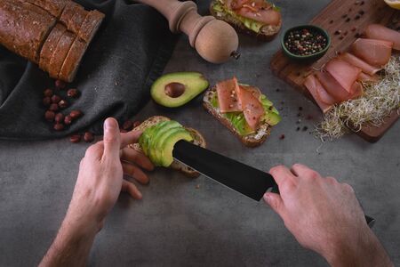 Chef making avocado sandwiches with bread, nuts, ham, sprouts and goat cheese on gray stone slate background. Man making breakfast in the kitchen. Healthy food preparation. Toned faded backgroundの写真素材