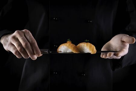 Sushi served on japanese knife in chef hands on dark background. Decorated salmon sashimi nigiri. Traditional japanese food. Copy space for textの写真素材