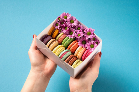 Woman holding in hands gift box with colorful french macaroons with flowers on bright blue background. Valentines Day (February 14), Mother's day, Birthday or Woman's day (8 March) concept. Copy spaceの写真素材