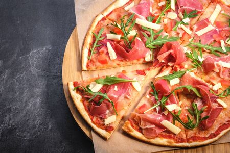 Pizza on wooden board on black stone background. Italian traditional food. Delicious pizza with serrano jamon, parmigiano-reggiano cheese and rucola. Restaurant menu. Copy space, top viewの写真素材
