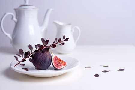 two figs on a white plate with a branch of barberry, white tea pot and cup on the table.の写真素材
