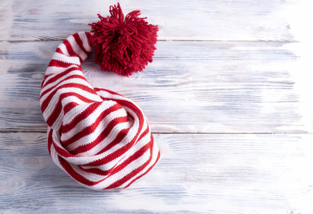One red and white striped cap with a long tail and a large red pompom is on a white wooden background, flat lay, top view.の写真素材