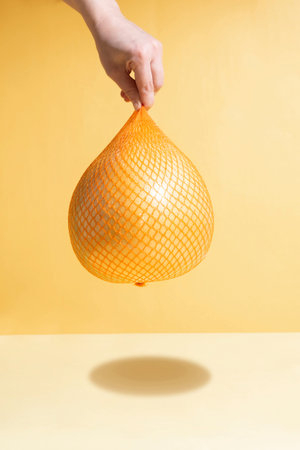 Female hand hangs yellow fresh pomelo in mesh packaging on a yellow and orange background in the...