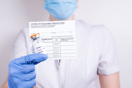 A woman doctor in a mask and gloves holding a vial with covid-19 vaccine and vaccination record...