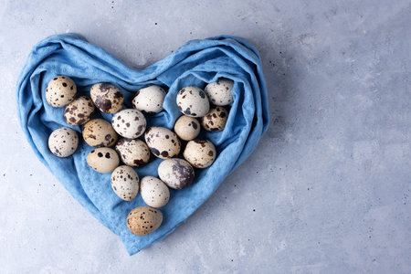 Quail eggs in a blue heart-shaped napkin on a gray background. Easter card, top view, copy space,...