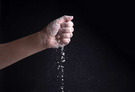 Female hand pouring flour on a black background, flying white powder, close up.の写真素材