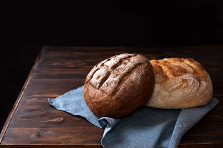 Round rye and wheat loaves of bread on a wooden table with a napkin on a black background, rustic style, close up.の写真素材