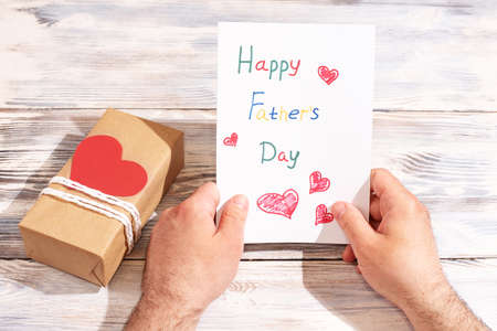 Male hands are holding a greeting card made by a child on a white wooden background with a gift, congratulations to father, close up.の写真素材