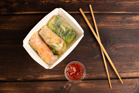 Three spring rolls in paper box with chopsticks and sauce on wooden background, asian fast food,...