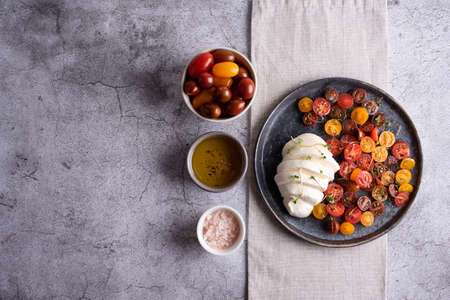 Caprese salad, plate with mozzarella ball and sliced colorful tomatoes, olive oil and vegetables on a gray background.の写真素材