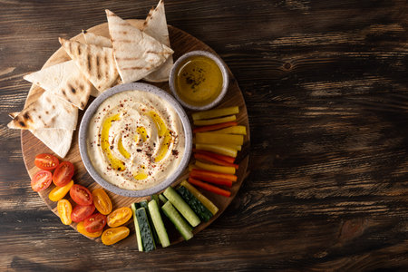 Sliced vegetables, pita and bowl with hummus made from chickpeas on dark wooden background, copy...