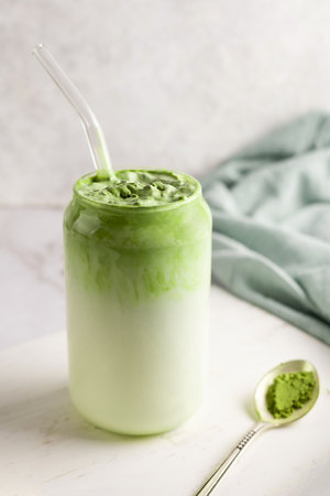 Glass of dalgona matcha latte and spoon of green tea powder on white background.の写真素材