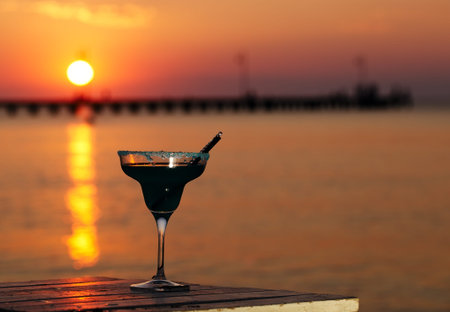 Glass of tropical cocktail standing on a wooden table overlooking a sunset ocean with the setting sun casting a fiery path across the waterの写真素材