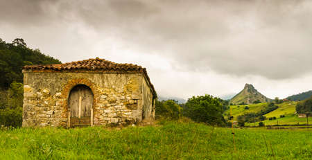 Asturian panoramic landscape with an old house in ruins and green slopes and mountains in the background with cloudy sky.の写真素材