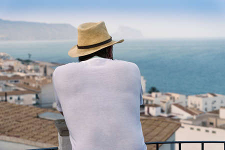 Man with his back turned and wearing a hat looking at a landscape of a coastal town with the sea in the background.のeditorial素材