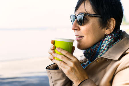 Mature woman wearing sunglasses drinking a hot beverage outdoors with blur background and copy space.の写真素材