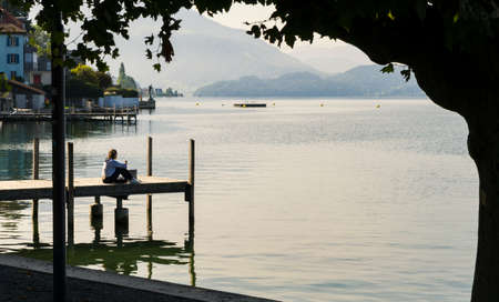 View of calm Lake Zugersee in the Swiss town of Zug with a small wooden harbor and a young girl sitting looking out over the lake at sunrise.のeditorial素材