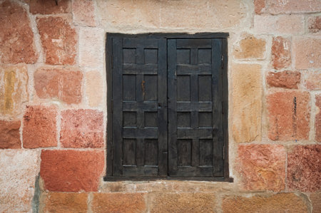 Detail of facade with stone wall in warm tones and a closed black wooden window in the center.
