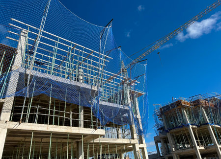 View of the construction site of an apartment building with cranes and safety nets at work, with blue sky and no people working.の写真素材