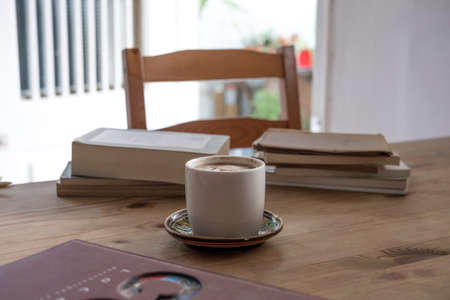 white cup with cappuccino coffee accompanied by a series of books on a rustic wooden tableの写真素材