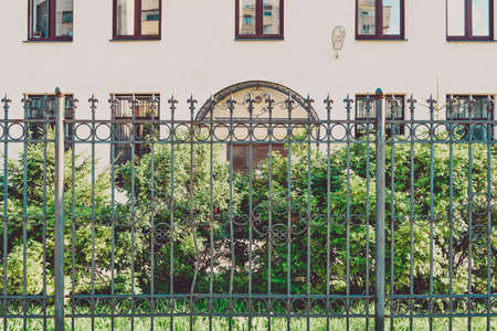 Generic urban city apartment building exterior with green bushes on front and steel decorative fence for street security. Old town house with light wall and small garden on summer sunny dayの写真素材