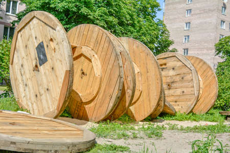 Large broken empty coils and reels with wooden cable drums, stacked on an open street area with sand and grass in the city park near to the housesの写真素材