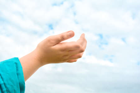 Close up of child hand in blue sleeve raised up over blue sky and clouds background. Children's hand reaches for the heaven. People, childhood, gesture and body parts conceptの写真素材
