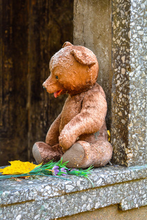 Weathered teddy bear toy with yellow maple leaf and flowers in opening of old crypt with child burial in cemetery in fall. The concept of sadness, grief over loss of child, deathの写真素材