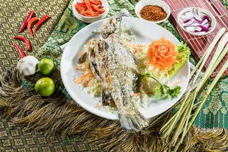 Thai-style grilled fish with crispy skin on shredded cabbage and carrot, surrounded by colorful spices and herbs.の写真素材
