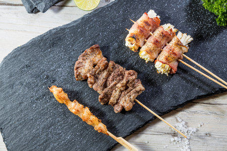 Grilled skewers with chicken, beef, and bacon-wrapped enoki mushrooms on slate.の写真素材