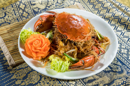 Whole crab served over glass noodles with vegetables and carved carrot garnish, styled on woven mat and patterned fabric.の写真素材