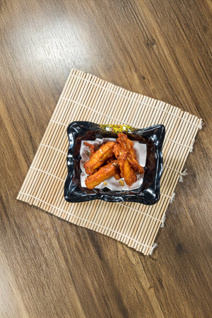 Glazed chicken wings coated in a sticky reddish-brown sauce, served on paper in a black dish atop a bamboo mat, highlighting sweet and spicy appeal.の写真素材