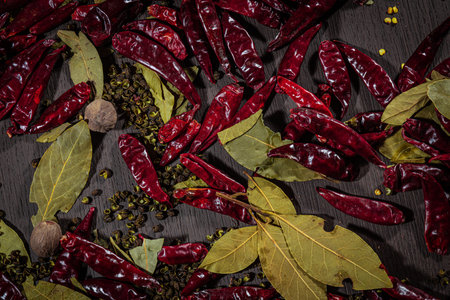 A rustic arrangement of dried red chilies, bay leaves, nutmeg, and Sichuan peppercorns on dark wood,...