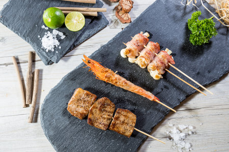 Grilled skewers with shrimp, meat cubes, and bacon-wrapped enoki mushrooms, garnished with lime and herbs.の写真素材