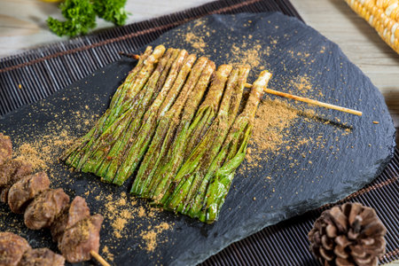 Grilled skewers of green onions and meat seasoned with dry spices, served on a black slate platter with parsley and pinecone on bamboo mat.の写真素材