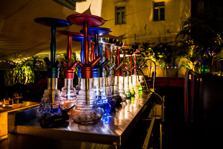 A row of colorful hookahs with glowing glass bases and metal stems arranged on a counter. Plants and...
