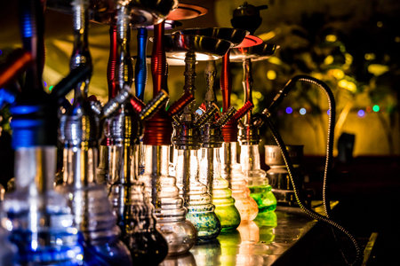 A row of colorful hookahs with coiled hoses and glowing bases on a reflective surface. Warm lighting...