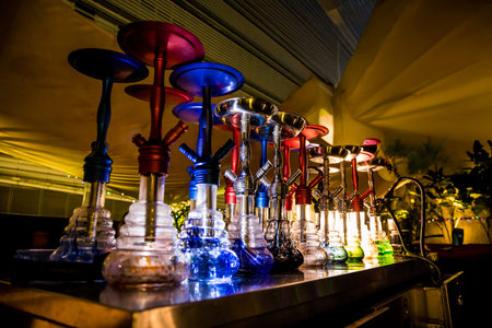 A row of colorful hookahs with distinct liquid bases and stems, set in a lounge with ambient...