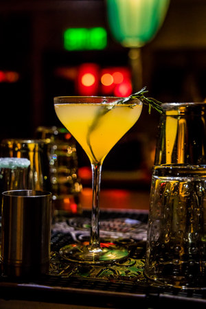 A yellow-orange cocktail in a martini glass garnished with rosemary. Bar tools and warm lighting create a festive and elegant lounge setting.の写真素材