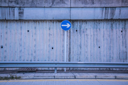 A blue circular traffic sign with a white arrow in front of a ribbed concrete wall, guardrail, and drainage grate.の写真素材