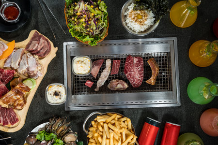 Korean BBQ setup with marbled beef, pork, seafood, rice, salad, sauces, fries, and colorful drinks around a central grill, vibrant and communal.の写真素材