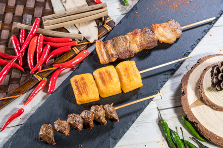 Three skewers with grilled pork belly, tofu cubes, and meat chunks, served with chili and rustic garnish.の写真素材