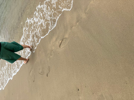 A person in green shorts walking barefoot along the shoreline, leaving footprints in the sand as...