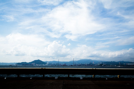 A waterfront road view of a coastal city with smokestacks, buildings, and green hills under a bright sky.の写真素材