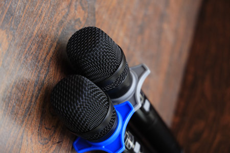 Two black handheld microphones with colored rings lie on a wooden surface, commonly used for events or public speaking.