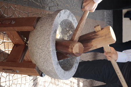 Two people pound steamed rice in a stone mortar using wooden mallets, one engraved with Korean text,...