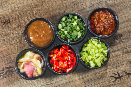 Six small bowls with Asian condiments including chili oil, hoisin sauce, green onion, sliced meat,...