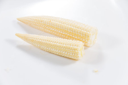 A high quality stock photo of two fresh yellow baby corn cobs with partial green husks on a white plate highlighting their crisp texture and natural color with a shallow depth of fieldの写真素材