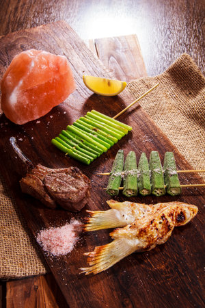 A rustic board with grilled lamb chop, fish, okra, asparagus, Himalayan salt and lemon wedge.の写真素材